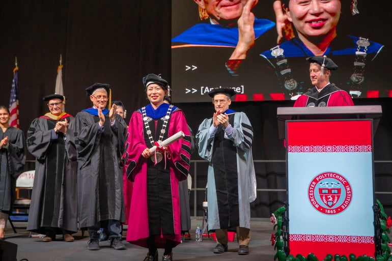A woman in a formal red robe is applauded by six other robed people standing on a stage.