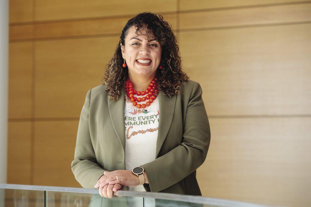 A woman with curly hair wearing a green blazer stands with her hands on a glass railing