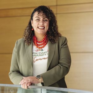 A woman with curly hair wearing a green blazer stands with her hands on a glass railing