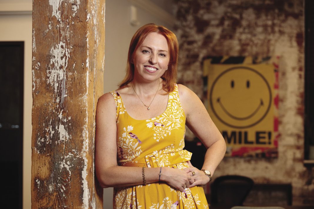 A woman leans against a wooden pillar while wearing a yellow dress.