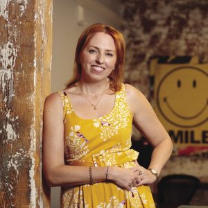 A woman leans against a wooden pillar while wearing a yellow dress.