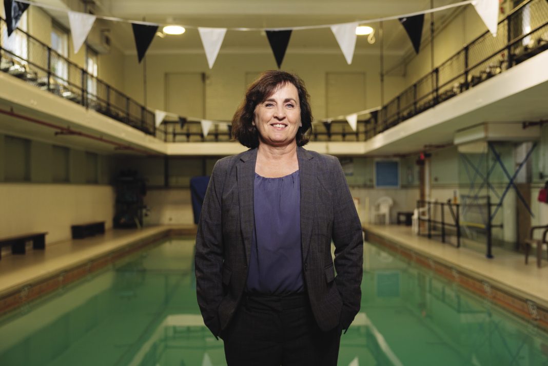 A woman wearing a grey suit stands in front of an indoor pool.