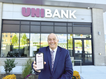 A man wearing a blue suit sits in front of a UniBank sign, holding his smartphone displaying a mobile banking app.