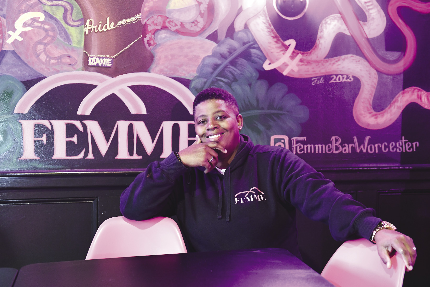 A woman with short hair sits at a table in front of a wall with a mural on it that reads 