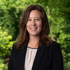 A woman with shoulder-length brown hair wears a black blazer and stand in front of greenery.