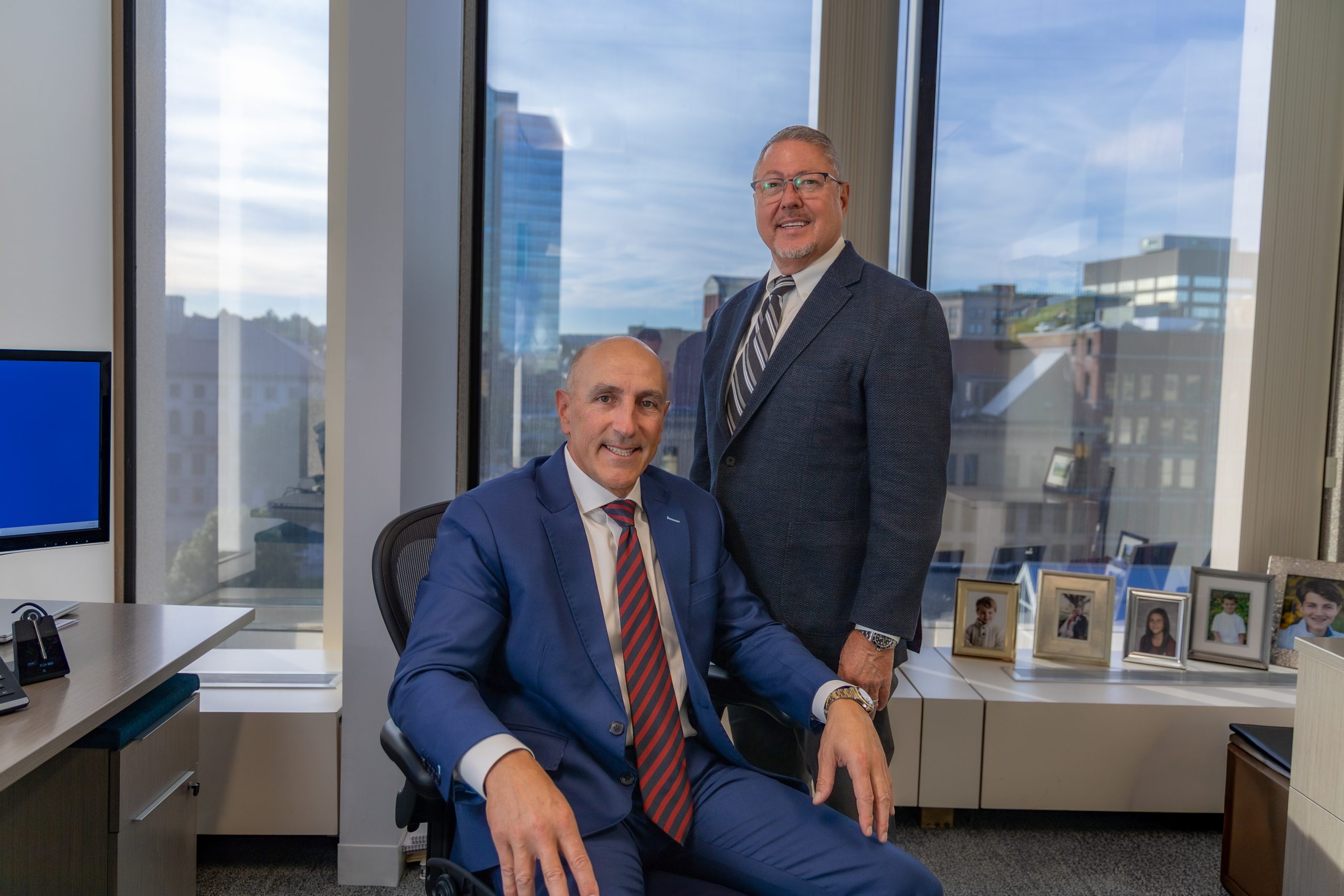 Two men in suits, one sitting while the other stands, in front of a window with a cityscape behind them
