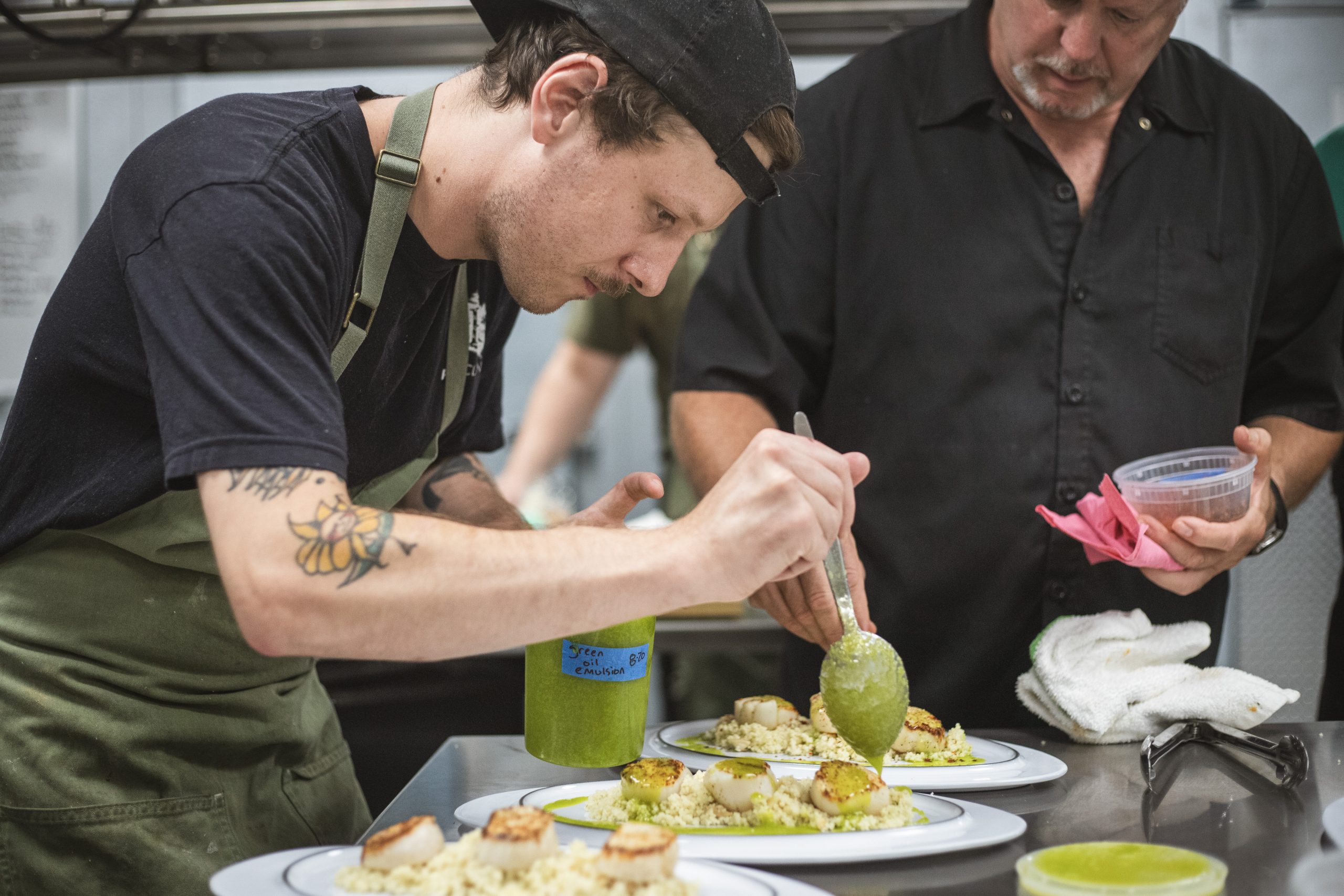 A chef drizzles a green sauce over a dish as another employee looks on