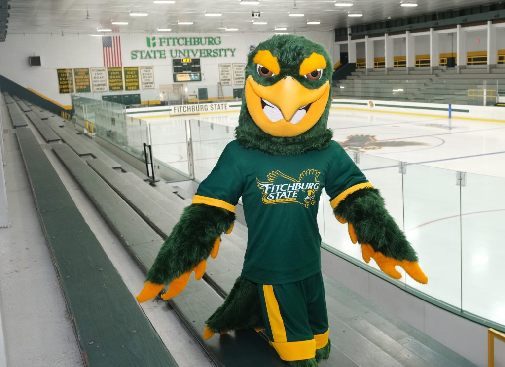 A falcon mascot wearing a Fitchburg State shirt stands in the stands of a hockey rink.