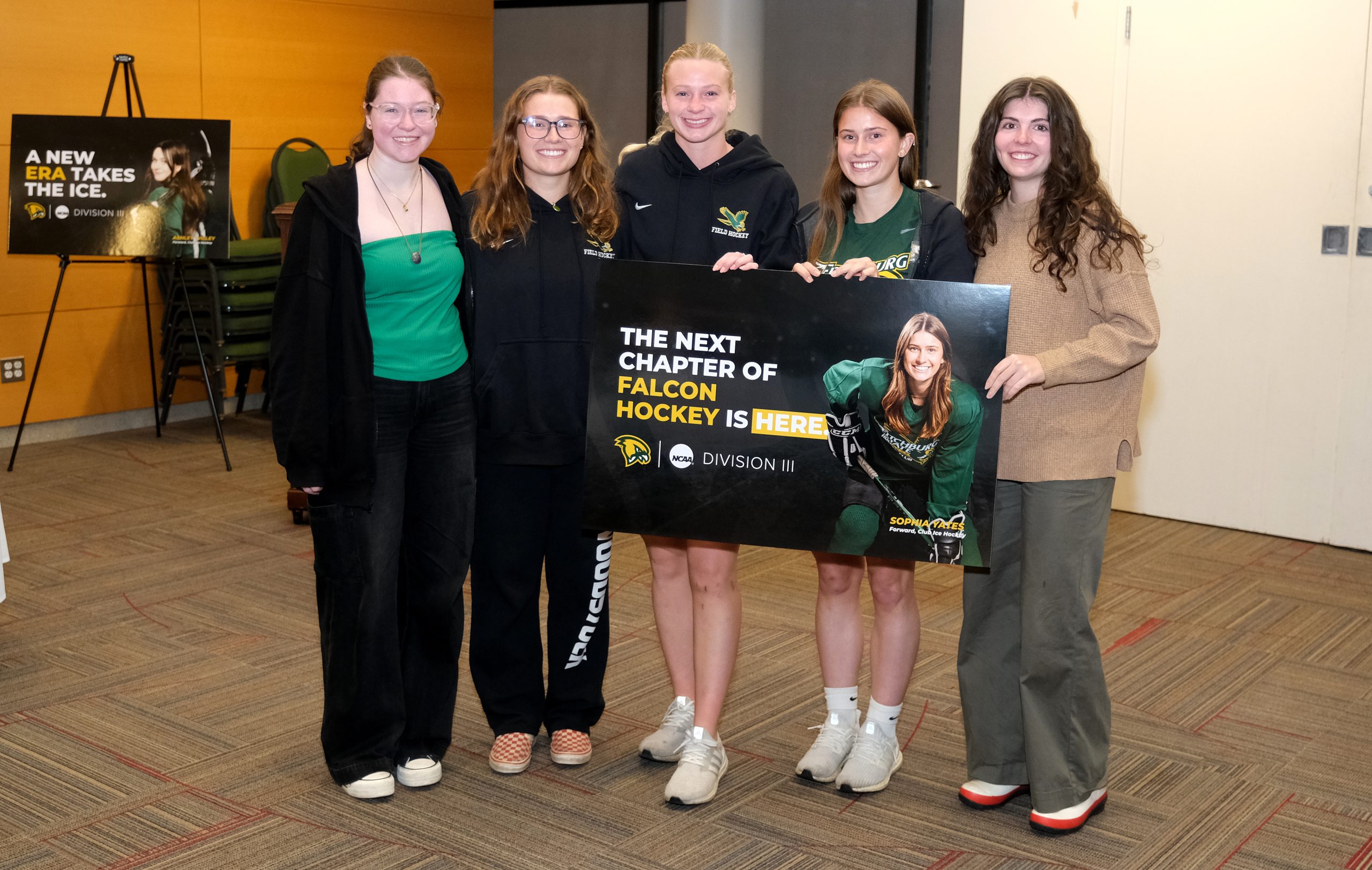Five young women standing inside hold a banner announcing the new women's hockey team at Fitchburg State