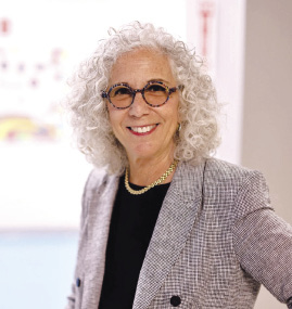 A woman with white curly hair wears glasses and a grey suit jacket.