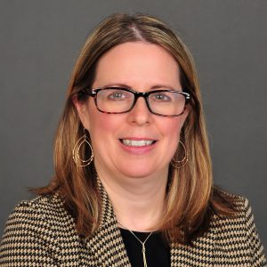 A woman with shoulder-length blonde hair wears a brown and black patterned blazer. 