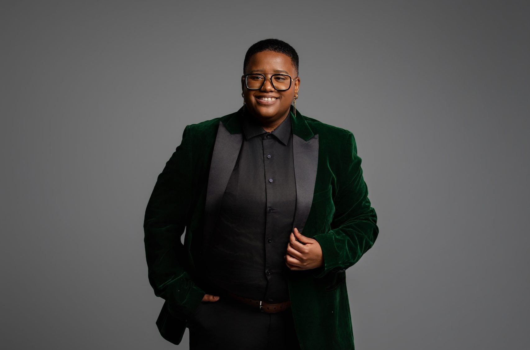 A woman in a dark green suit poses for a photo in front of a grey backround