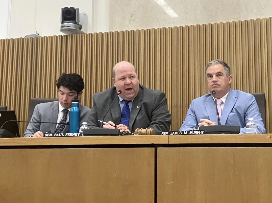 Three men in suits sit at a panel during a hearing