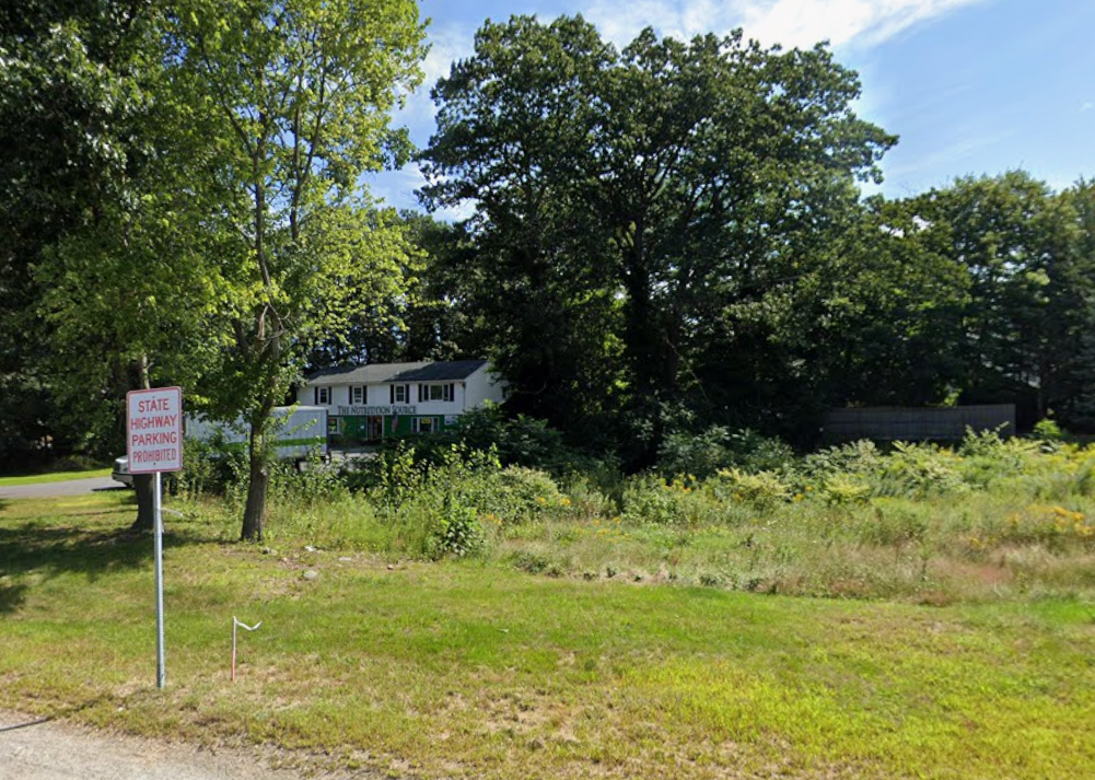 An overgrown field with a two-story house behind trees