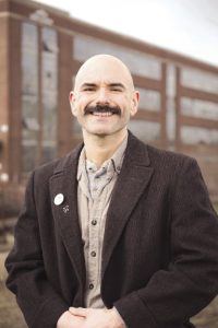 A man with a mustache wears a brown suit jacket.