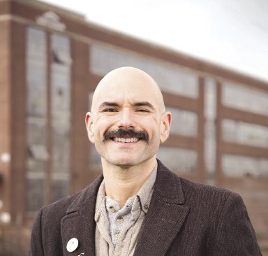 A man with a mustache wears a brown suit jacket.