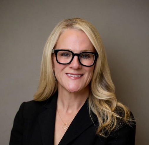 A woman with long, wavy blonde hair wears black glasses and a black blazer.
