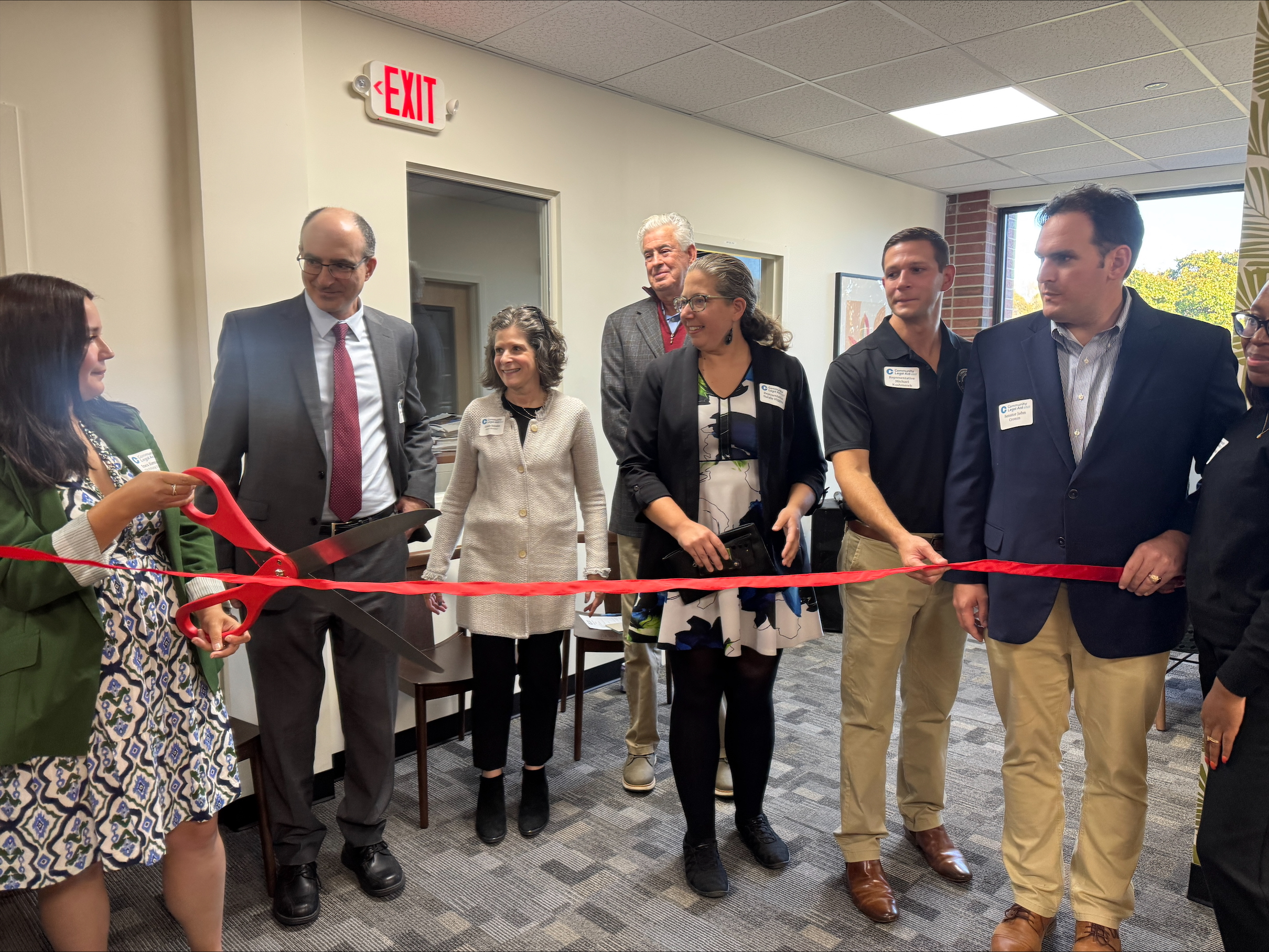 A group of people stand in a line inside an office with one person cutting a red ribbon with a pair of large scissors.