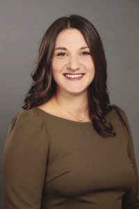 A woman with long brown hair wears a dark greenish brown top.