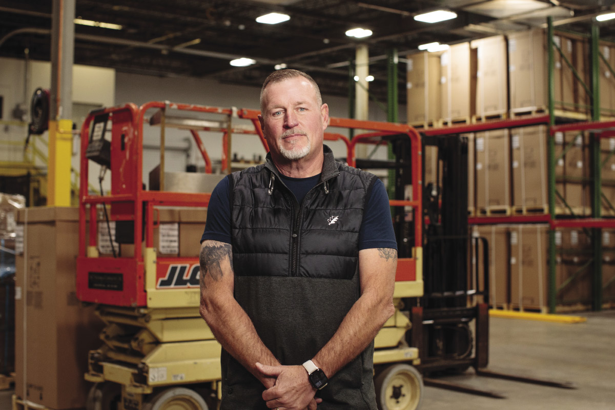 Man with tattos wearing a black vest smiles with his hands folded in front of a forklift in an industrial building.