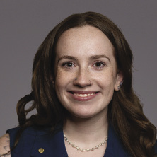 A woman with long brown hair wears a dark blue vest and a pearl necklace.