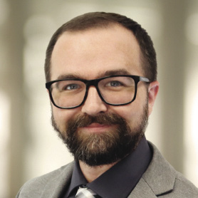 A man with short dark hair and a goatee wears a grey suit jacket.