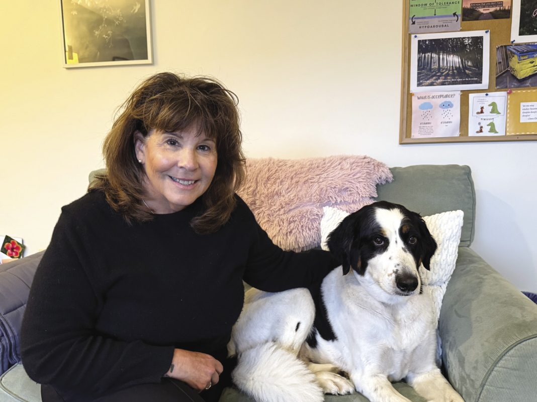 A woman with brown hair and a black sweater sits on a couch with a black and white dog.