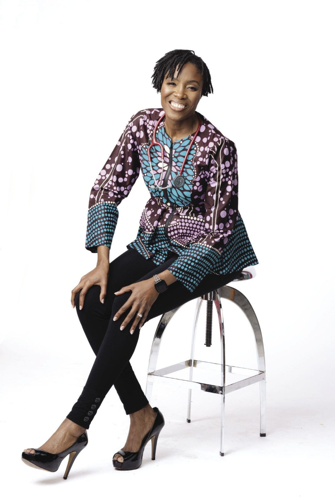 A woman with short dark hair wears a colorful blouse and black pants while sitting on a stool.