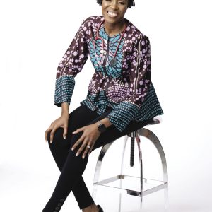A woman with short dark hair wears a colorful blouse and black pants while sitting on a stool.