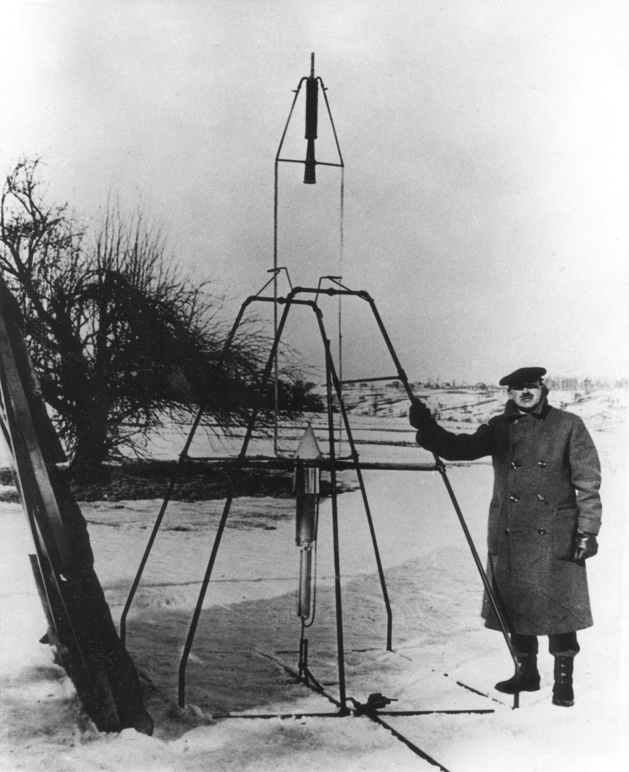A black-and-white photo of a man standing next to a small rocket at the top of a metal support structure