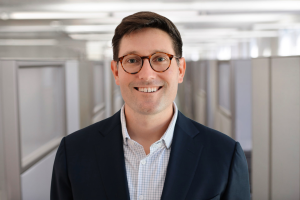 Man in suit with classes standing in front of backdrop featuring office cubicles. 