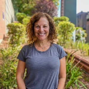 A woman in a gray shirts stands in some greenery.
