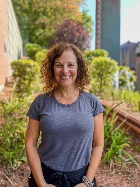 A woman in a gray shirts stands in some greenery.