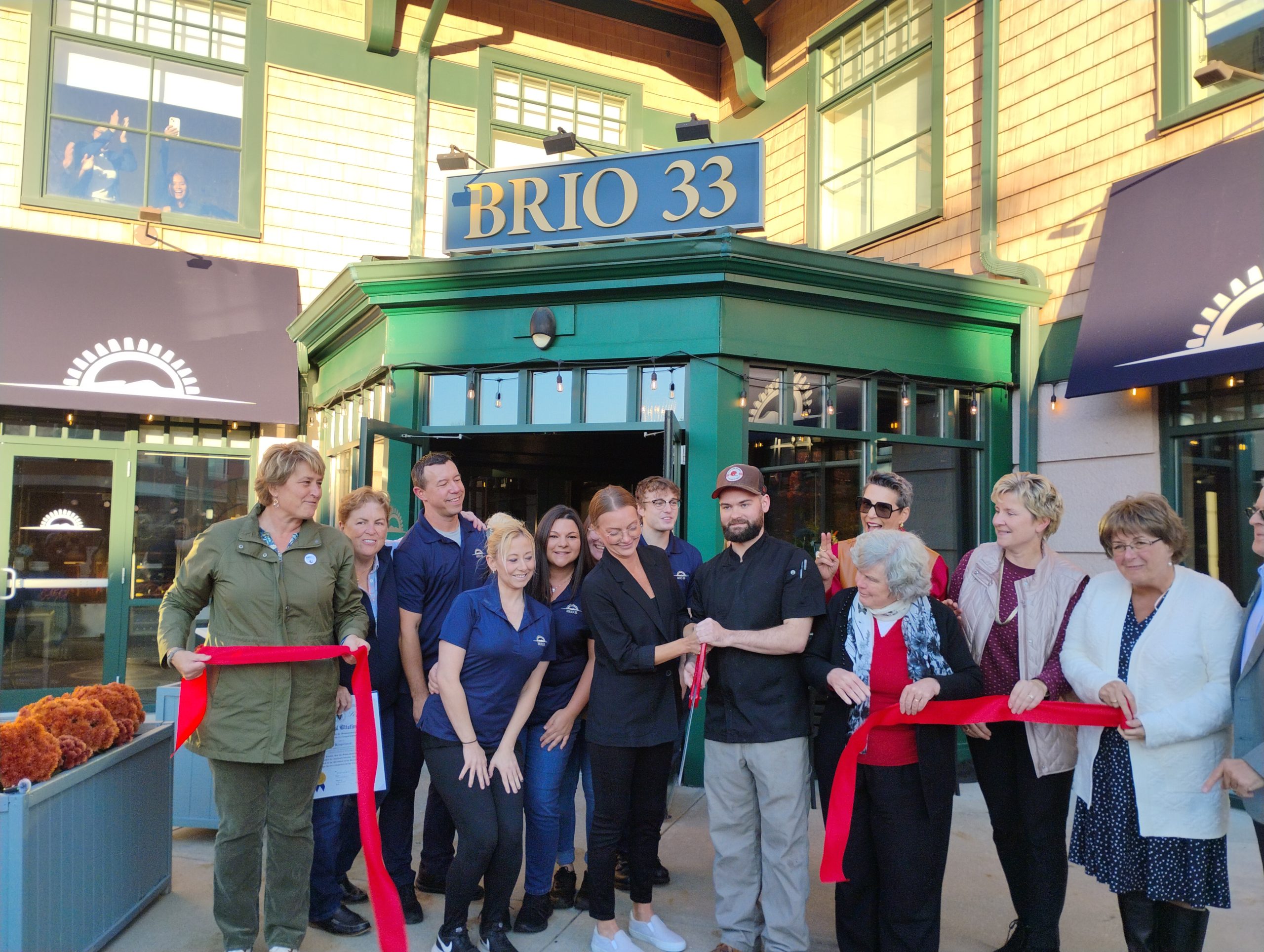 A group of people gather in front of a restaurant for a ribbon cutting ceremony, as three people look on from a second story window behind them.
