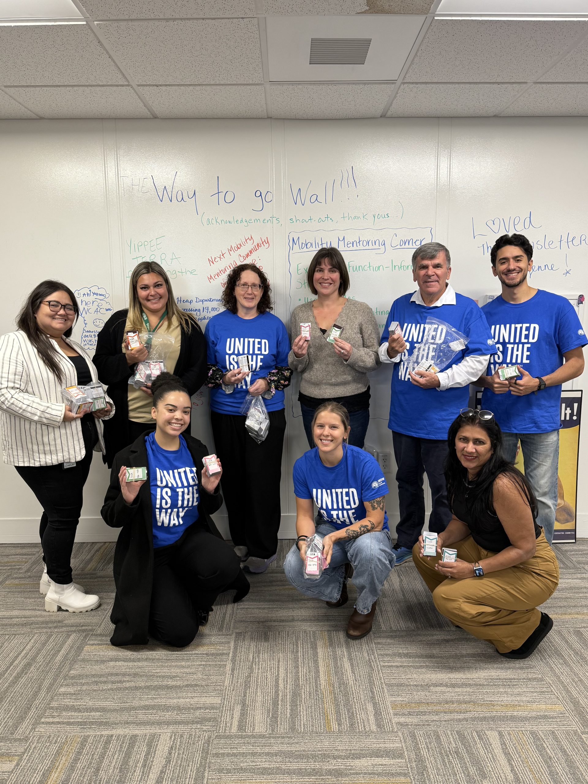 A group of people hold stacks of gift cards while standing in front of a white board.