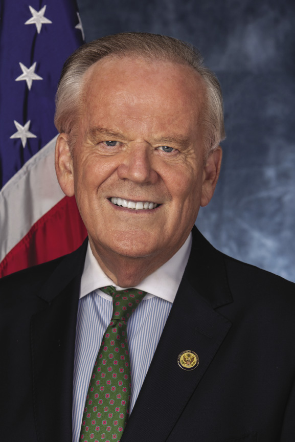 A man with grey hair wears a black suit jacket, blue button down, and green tie.