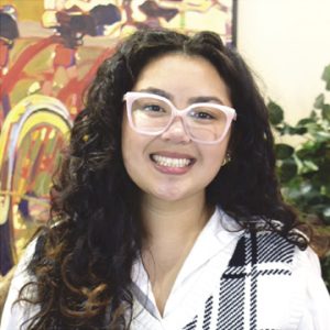 A woman with long, dark curly hair wears a black and white top and light pink glasses.