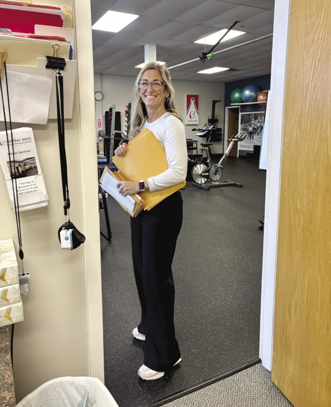 A woman with long blonde hair stands holding several manila folders.