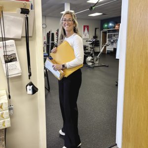 A woman with long blonde hair stands holding several manila folders.