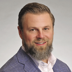 A man with a mustache and beard wears a light blue blazer and white button down.