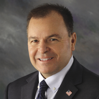 A man wears a black suit jacket with an American flag pin on it, a white button down, and a black tie.
