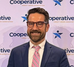A man with dark hair wears glasses, a dark blue suit jacket, white button down shirt, and pink tie.