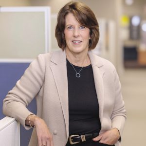 A woman with a light brown bob wears a tan blazer, a black top, and black pants