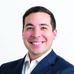 A man with short dark hair wears a blue suit jacket and white button down.