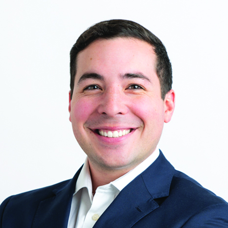 A man with short dark hair wears a blue suit jacket and white button down.