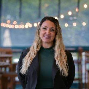 A woman with long blonde hair wears a black blazer and dark green mock-neck turtleneck.
