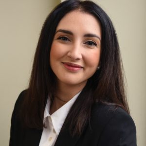 A headshot of a woman with long dark brown hair, wearing a black blazer and a white button down shirt