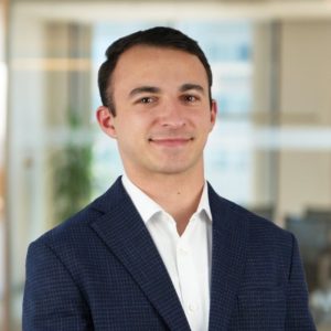 A headshot of a man with dark brown hair, wearing a dark blue blazer and a white button down.