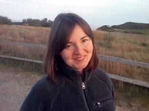 A woman with short brown hair wears a blue zip-up sweater while standing in front of a field.