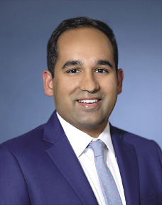 A man wears a dark blue blazer, a white button down shirt, and a light blue tie.